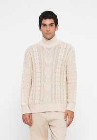 Hombre con suéter de punto beige con cuello alto y pantalones beige claros de pie frente a un fondo blanco, mirando hacia adelante.