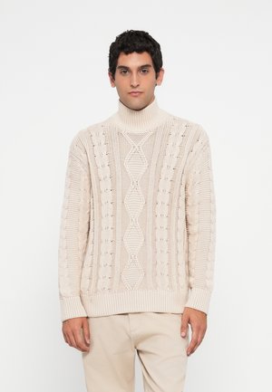 Man in beige cable-knit turtleneck sweater and light beige pants standing against a white background, facing forward.