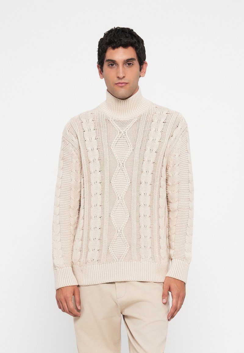 Hombre con suéter de punto beige con cuello alto y pantalones beige claros de pie frente a un fondo blanco, mirando hacia adelante.