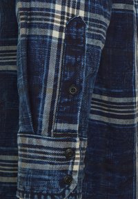 Sinivalkoruudullinen paita, jossa on pehmeä, teksturoidu kangas, napillinen hihansuu ja kontrastin luovat mustat napit. Suuri ruutukuvio.