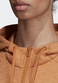 Sweat à capuche zippé en tissu texturé orange. Comprend une capuche, des cordons de serrage et une fermeture éclair visible. Gros plan sur la zone du cou.