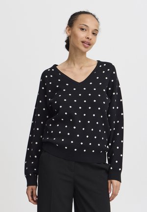 Schwarzer V-Ausschnitt-Pullover mit weißen Punkten, langen Ärmeln und gerippten Bündchen, kombiniert mit schwarzen Hosen. Weicher Stoff, lässige Passform.