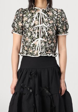 Vrouw draagt een zwarte bloemige korte mouw blouse met witte randafwerking en strikjes aan de voorkant, gecombineerd met een zwarte rok met lintdetails.