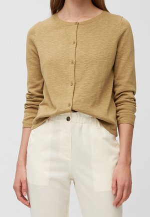 Person wearing a light brown buttoned cardigan with long sleeves and high-waisted off-white trousers against a neutral background.