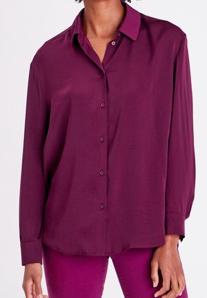 Femme portant une chemise prune ample à manches longues boutonnée, associée à un pantalon magenta, debout devant un fond uni.