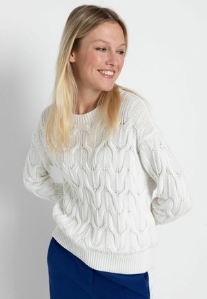 Weißer Strickpullover mit einem strukturierten Wellenmuster, rundem Halsausschnitt und geripptem Saum. Das Model kombiniert ihn mit dunkelblauen Hosen.