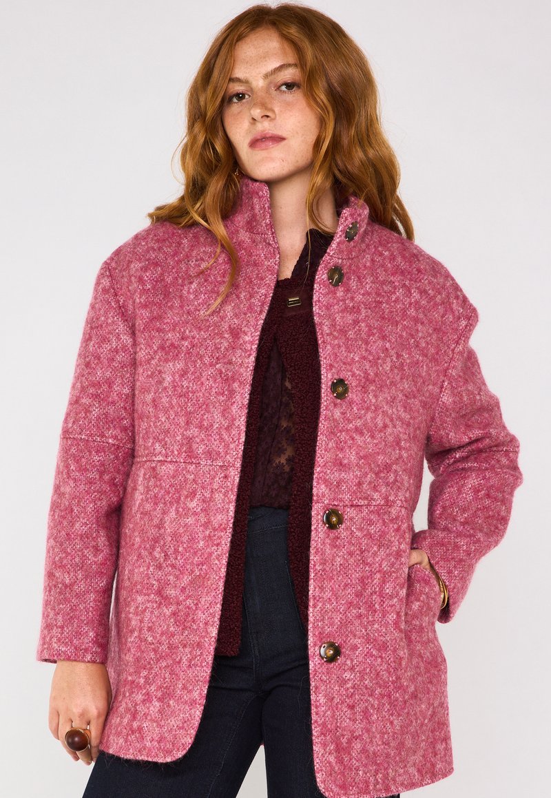 Cappotto in misto lana rosa con finitura texturizzata; presenta un colletto alto, tre bottoni e tasche laterali. Il design è dritto e leggermente oversize.