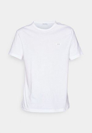 T-shirt en coton blanc à manches courtes, col rond et un petit label de la marque sur la poitrine. Texture lisse et design minimaliste.