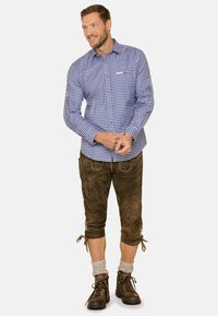 Blau-weiß kariertes Hemd mit langen Ärmeln, kombiniert mit braunen knielangen Shorts und braunen Wanderschuhen. Strukturiertest Gewebe mit Knöpfen.