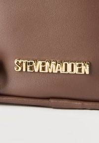 Κοντινή εικόνα καφέ δερμάτινης επιφάνειας με το χρυσό λογότυπο της μάρκας "Steven Madden" προσαρτημένο.