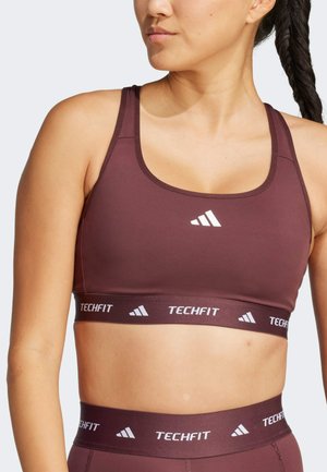 Sujetador deportivo burdeos con escote redondeado y tirantes ajustables. Presenta un logo TECHFIT en blanco y una banda de contraste con detalles del logo.