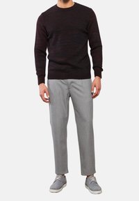 Un uomo che indossa un maglione scuro a fantasia, pantaloni cropped grigi chiari e scarpe casual grigie con lacci su uno sfondo bianco.