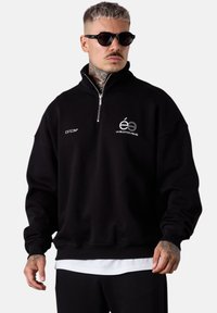 Homme avec des tatouages et des lunettes de soleil portant un sweat à capuche noir zippé avec des logos en texte blanc et une chemise blanche qui dépasse en dessous.