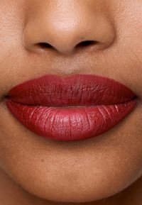 Matowa, głęboka czerwona szminka na pełnych ustach, podkreślająca teksturę i wyraźny kształt. Naturalny odcień skóry w tle uzupełnia kolor.