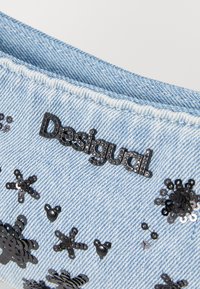 Denim w jasnoniebieskim kolorze ma czarne logo „Desigual” oraz kilka dekoracyjnych czarnych cekinów w kształcie kwiatów i abstrakcyjnym.