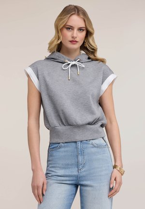 Blonde Frau trägt ärmellosen grauen Hoodie mit weißen Kordeln und hellblauen hoch taillierten Jeans, steht vor neutralem Hintergrund.