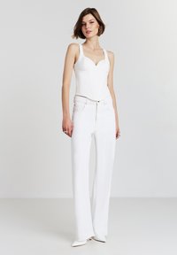 Una donna è in piedi contro uno sfondo semplice indossando un top aderente bianco senza maniche, jeans bianchi a vita alta, scarpe bianche con punta e anelli d'argento.