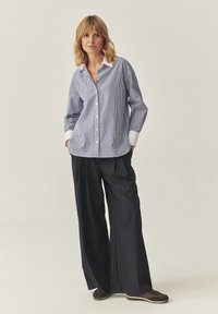 Camicia a righe blu e bianche con colletto bianco, abbinata a pantaloni a gamba larga neri con righe sottili e scarpe da ginnastica scure.