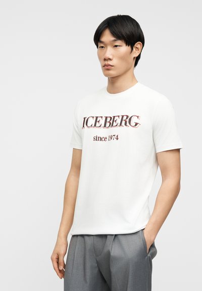 Νέος άνδρας που φοράει λευκό Τ-shirt με το κείμενο "ICEBERG since 1974", γκρι παντελόνι, στέκεται με το ένα χέρι στην τσέπη μπροστά σε απλό φόντο.