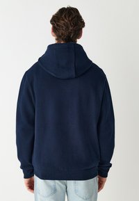 Laivastonsininen huppari, jossa on ribbineulostekstuuri, iso huppu ja joustavat hihansuut. Päällä vaaleansiniset farkut, jotka korostavat rentoa tyyliä.