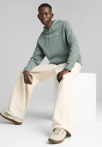 Hætte sweatshirt i dæmpet grøn, parret med off-white brede bukser og beige sneakers. Har logo på brystet og afslappet pasform.