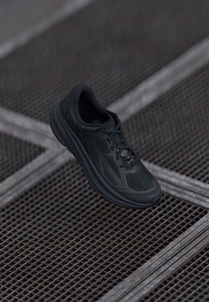 Czarny but sportowy z siateczkową górą i sznurowadłami, zawieszony nad metalową kratownicą z przecinającymi się metalowymi paskami.