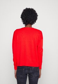 Jasnoczerwony dziany sweter o luźnym kroju i opuszczonych ramionach. Prążkowany dekolt i mankiety, w połączeniu z ciemnymi dżinsami, widok od tyłu.