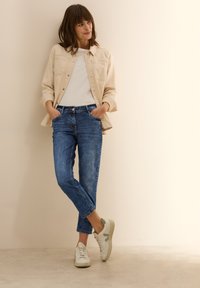 Ljusbeige denimjacka över en vit T-shirt, tillsammans med blå jeans. Skorna är ljusgrå sneakers med en subtil accent.