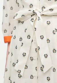 Κρέμ φόρεμα με μαύρα και πορτοκαλί λουλουδένια σχέδια, κουμπιά μπροστά, εφαρμοστή μέση με υφασμάτινη ζώνη δεμένη, και μανσέτα με πορτοκαλί περιθώριο.