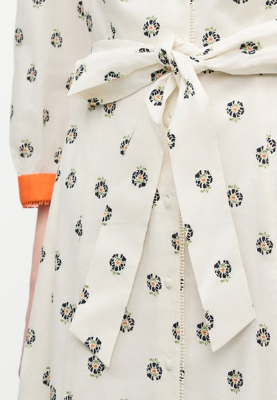 Robe crème à imprimés floraux noirs et orange, boutons sur le devant, taille cintrée avec une ceinture en tissu nouée, et poignet de manche bordé d'orange.