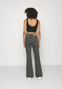 Top corto negro combinado con jeans oscuros grises de cintura alta y acampanados, con una etiqueta de cuero marrón y bolsillos traseros visibles. Zapatillas blancas completan el look.