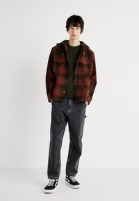 Camisa de franela a cuadros rojiblancos con capucha, llevada sobre una camiseta verde oscura, combinada con pantalones cargo gris oscuro y zapatillas Vans negras.