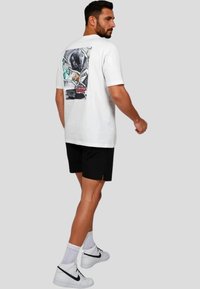 Hvid bomulds T-shirt med et farverigt grafik på ryggen. Båret sammen med sorte shorts og hvide højhalsa sneakers.