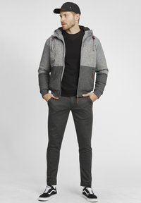 Gray and black bomber jacket with hood, textured fabric, zip front, gray sweater, dark pants, and black sneakers with white accents.