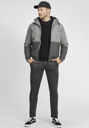 Sivá a čierna bomber bunda s kapucňou, textúrovaná látka, zips vpredu, sivý sveter, tmavé nohavice a čierne tenisky s bielymi detailmi.