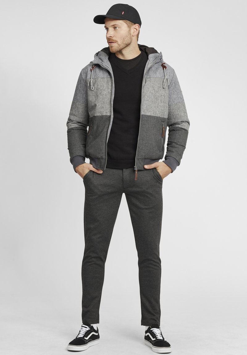 Gray and black bomber jacket with hood, textured fabric, zip front, gray sweater, dark pants, and black sneakers with white accents.
