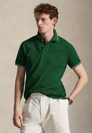 Ung mand med krøllet hår iført en mørkegrøn polo og hvide bukser, der kigger til siden med den ene hånd i lommen.