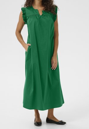 Vrouw in een mouwloze groene jurk tot de enkels met gerimpelde schouders en zwarte ballerina's, staande met één hand in de zak.