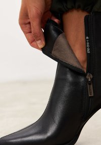 Bottine en cuir noir avec une finition lisse et un détail de fermeture éclair. L'intérieur est doublé d'un matériau gris doux pour plus de confort.