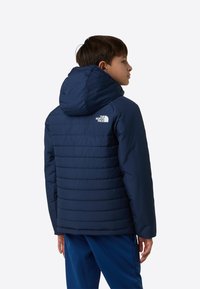 Tummansininen toppatakki hupulla, jossa on pystysuora tikattu kuvio ja The North Face -logo vasemmassa olkapäässä.