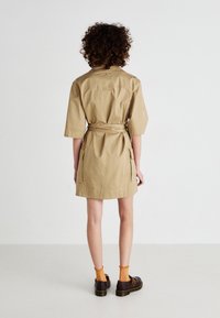 Khaki sukienka na krótki rękaw z paskiem, krótkimi rękawami oraz bocznymi kieszeniami. Materiał jest gładki i lekki, w połączeniu z ciemnymi butami i pomarańczowymi skarpetkami.