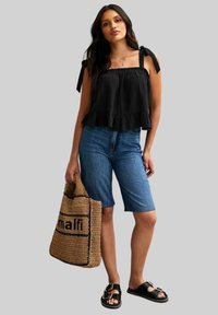 Svart strukturerad topp med volangkant, denimbermuda-shorts, vävd tote-väska med svart text och svarta sandaler med spänne.