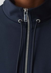 Mörkblå zip-up jacka med hög krage, silverdetaljer och elastiska dragsko. Tillverkad av mjukt, lätt tyg med en figurnära design.