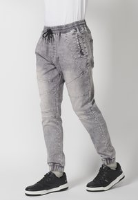 Šedé joggingové kalhoty s elastickým pasem, šňůrkou na stažení, bočními zipovými kapsami a prošitými panely. Nosené s černými teniskami.