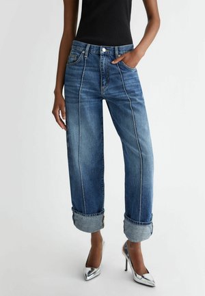 Person iført brede, opsmøgede blå jeans, en sort ærmeløs top og skinnende sølvfarvede stiletter, står med den ene hånd i lommen.