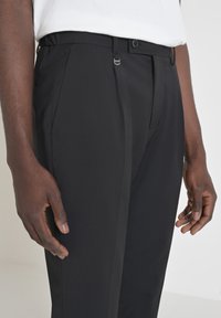 Zwarte maatwerk pantalons hebben een gladde textuur, plooien aan de voorkant en een stevige sluiting. De tailleband heeft riemlussen en een knoop.