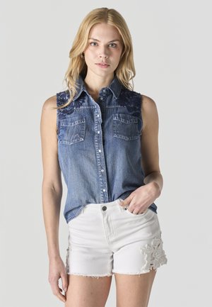 Mujer rubia con camisa de mezclilla azul sin mangas abotonada y pantalones cortos blancos con detalle de encaje, de pie con la mano en el bolsillo.