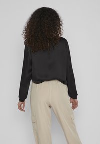 Camicia nera in satin a maniche lunghe con polsini arricciati, abbinata a pantaloni elasticizzati beige chiaro con vita elasticizzata e tasche laterali.