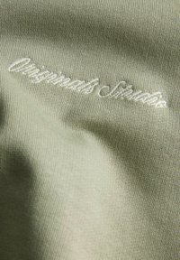 Svetlo zelena tkanina s teksturiranim pletenim vzorcem in subtelnim leskom. Vsebuje belo izšito besedilo "Originals Store."