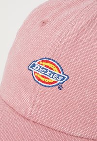 Roza kapa s teksturirano tkanino, z vezenim logotipom Dickies v modri, rumeni in rdeči barvi. Oblikovanje vključuje ukrivljen vizir.
